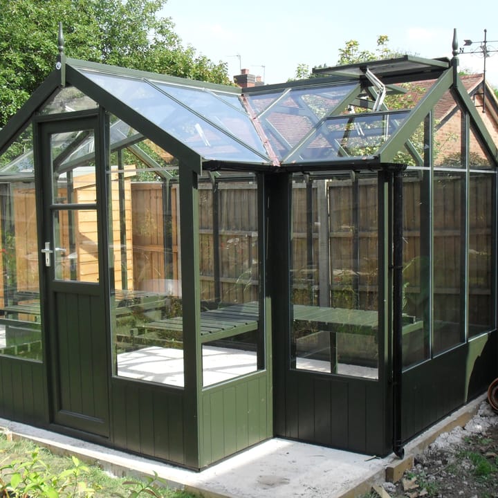 6ft 8in deep x 11ft 5in wide Swallow Cygnet t-shaped greenhouse. The Cygnet incudes front returned staging which sits at the front of the greenhouse and wraps around into the porch area. Optional 'Olive' painted finish has been added. The Cygnet can be constructed in either dwarf wall format or as pictured here freestanding. There is no extra cost for the dwarf wall option.