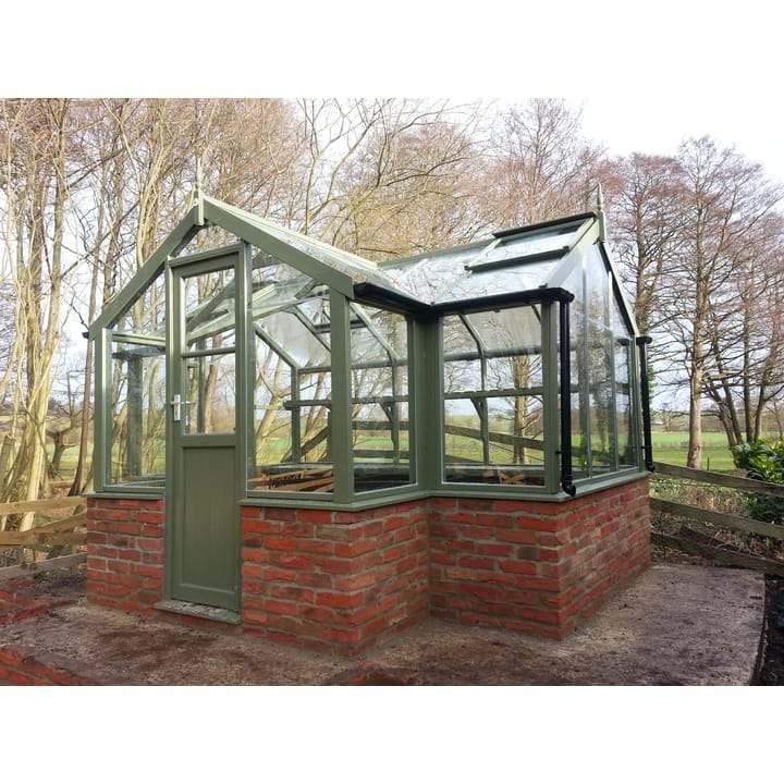 6ft 8in deep x 11ft 5in wide Swallow Cygnet t-shaped greenhouse. The Cygnet incudes front returned staging which sits at the front of the greenhouse and wraps around into the porch area. Optional 'Bracken' painted finish has been added as well as high level shelving and staging to the rear. The Cygnet can be constructed in either freestanding dwarf wall format or as pictured here dwarf wall. There is no extra cost for the dwarf wall option.