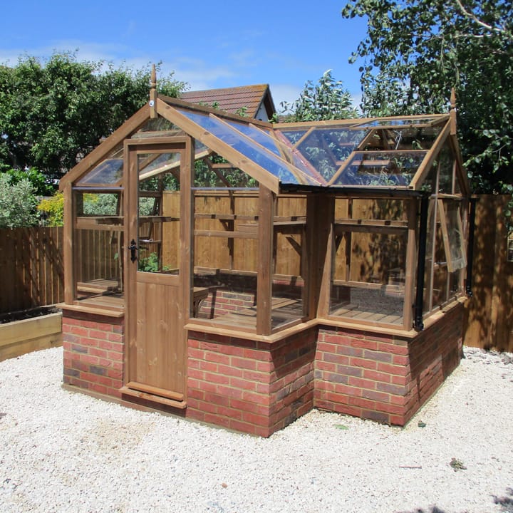 6ft 8in deep x 11ft 5in wide Swallow Cygnet t-shaped greenhouse. The Cygnet incudes front returned staging which sits at the front of the greenhouse and wraps around into the porch area. Optional front returned high level shelving and high level shelving to the rear has been added. The Cygnet can be constructed in either freestanding dwarf wall format or as pictured here dwarf wall. There is no extra cost for the dwarf wall option.