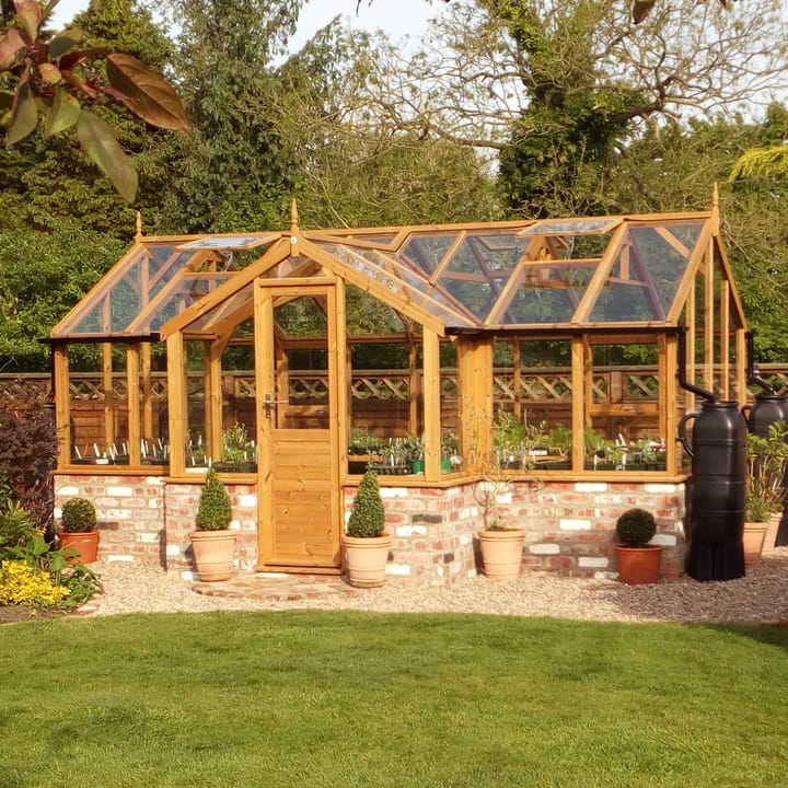 8ft 9in deep x 15ft 10in wide Swallow Mallard t-shaped greenhouse. The Mallard incudes front returned staging which sits at the front of the greenhouse and wraps around into the porch area. Optional 'Oiled finish' has been applied to the Thermowood timber.  The Mallard can be constructed either freestanding or as pictured here onto a dwarf wall. There is no extra cost for the dwarf wall option.