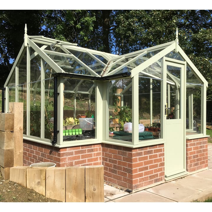 8ft 9in deep x 11ft 5in wide Swallow Mallard t-shaped greenhouse. The Mallard incudes front returned staging which sits at the front of the greenhouse and wraps around into the porch area. Optional 'Vert de terre painted finish' has been applied to the Thermowood timber.  The Mallard can be constructed either freestanding or as pictured here onto a dwarf wall. There is no extra cost for the dwarf wall option.
