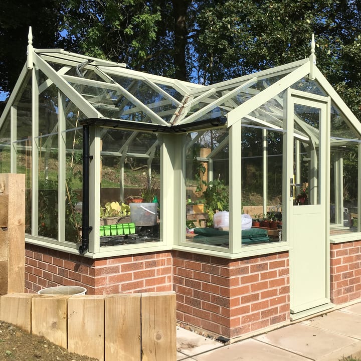 8ft 9in deep x 11ft 5in wide Swallow Mallard t-shaped greenhouse. The Mallard incudes front returned staging which sits at the front of the greenhouse and wraps around into the porch area. Optional 'Vert de terre painted finish' has been applied to the Thermowood timber.  The Mallard can be constructed either freestanding or as pictured here onto a dwarf wall. There is no extra cost for the dwarf wall option.