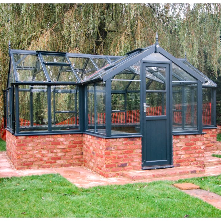 8ft 9in deep x 15ft 9in wide Swallow Mallard t-shaped greenhouse. The Mallard incudes front returned staging which sits at the front of the greenhouse and wraps around into the porch area. Optional 'Anthracite painted finish' has been applied to the Thermowood timber.  The Mallard can be constructed either freestanding or as pictured here onto a dwarf wall. There is no extra cost for the dwarf wall option.