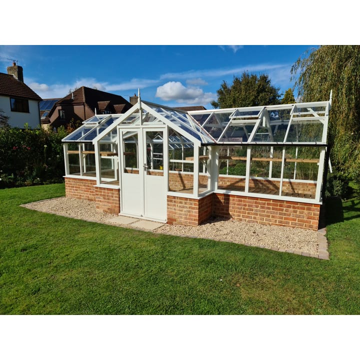 8ft 9in deep x 21ft 1in wide Swallow Swan t-shaped greenhouse. The Swan incudes front returned staging which sits at the front of the greenhouse and wraps around into the porch area. Optional 'Lily White' painted finish has been applied, as has front returned high level shelving. The Swan can be constructed either freestanding or as pictured here in dwarf wall format. There is no extra cost for the dwarf wall option.
