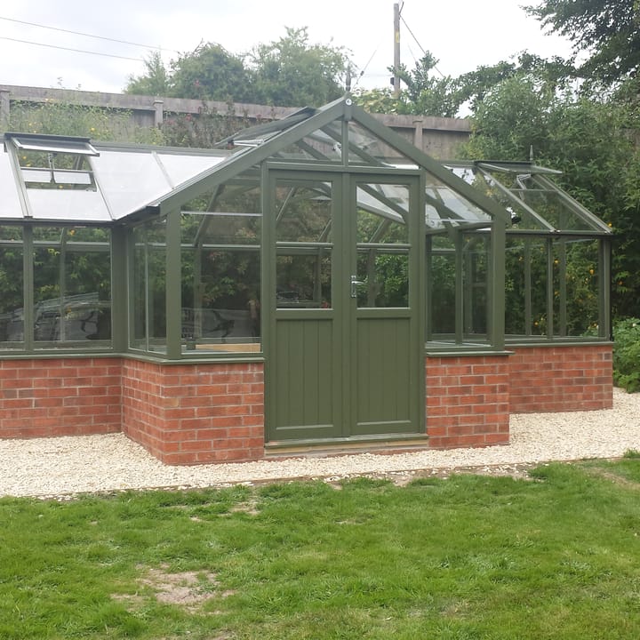 8ft 9in deep x 21ft 1in wide Swallow Swan t-shaped greenhouse. The Swan incudes front returned staging which sits at the front of the greenhouse and wraps around into the porch area. Optional 'Bracken' painted finish has been applied. The Swan can be constructed either freestanding or as pictured here in dwarf wall format. There is no extra cost for the dwarf wall option.