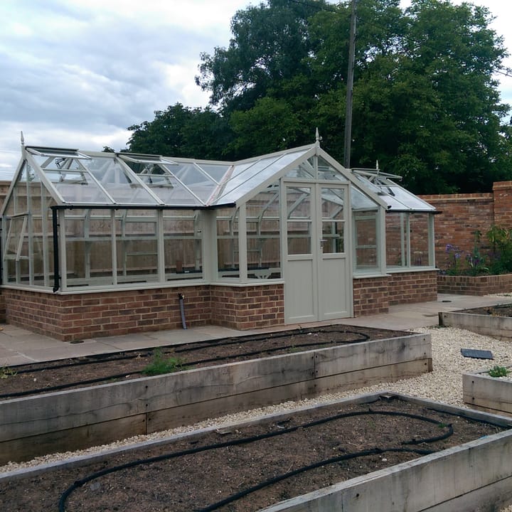 8ft 9in deep x 21ft 1in wide Swallow Swan t-shaped greenhouse. The Swan incudes front returned staging which sits at the front of the greenhouse and wraps around into the porch area. Optional 'Vert de Terre' painted finish has been applied, as has additional staging and high level staging to the rear. The Swan can be constructed either freestanding or as pictured here in dwarf wall format. There is no extra cost for the dwarf wall option.
