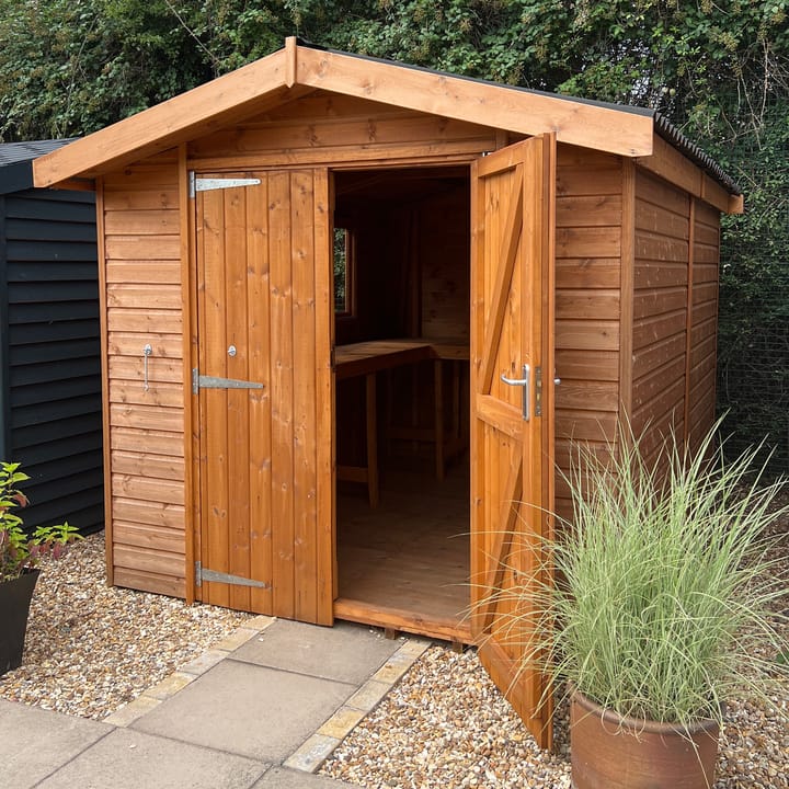 This 8ft x 10ft Heavy Duty Apex is constructed in heavy duty redwood cladding. A roof overhang and opening windows are standard features. Ironmongery is available in a choice of black or as pictured here chrome. Optional double doors, onduline roof and a workbench have been added.